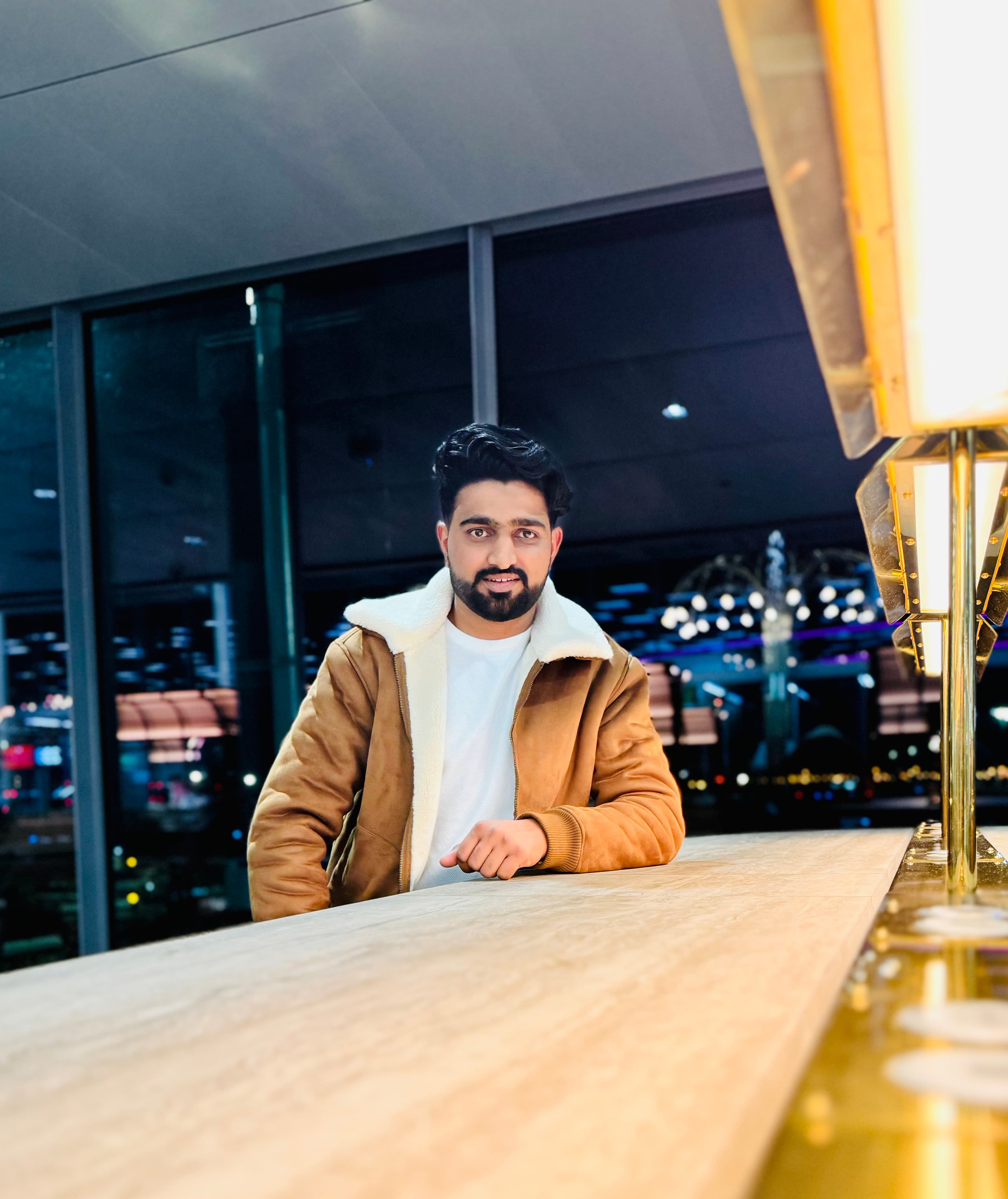 Bearded man in a brown shearling jacket leaning on a wooden counter at night.